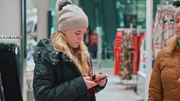 年轻迷人的女孩看着智能手机她在服装店商场里视频的预览图