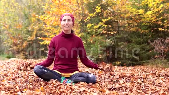 一个年轻的女孩坐在森林的空地上兴高采烈地抛出黄色的叶子视频的预览图