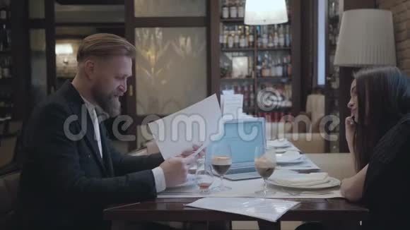 布鲁内特女士和金发胡须男坐在咖啡馆的桌子旁工作和喝啤酒笔记本电脑报纸和视频的预览图