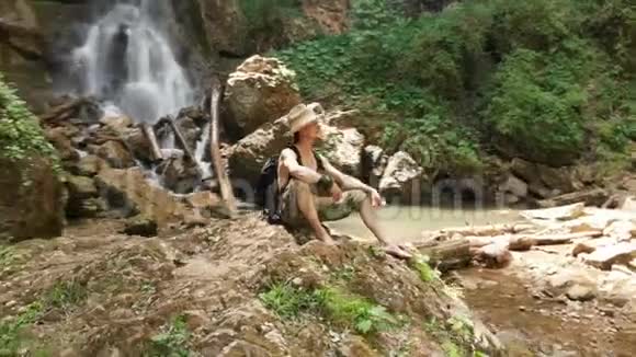 一位戴着草帽和背包的男性旅行者坐在岩石上观看雄伟的瀑布视频的预览图