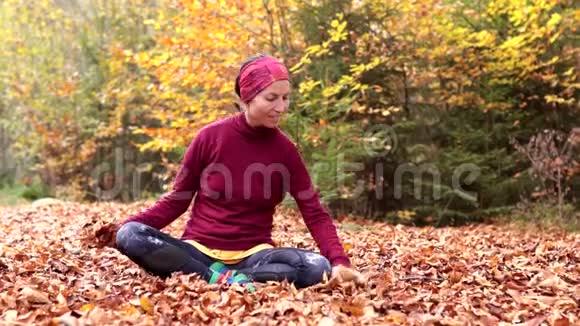 一个年轻的女孩坐在森林的空地上兴高采烈地抛出黄色的叶子视频的预览图