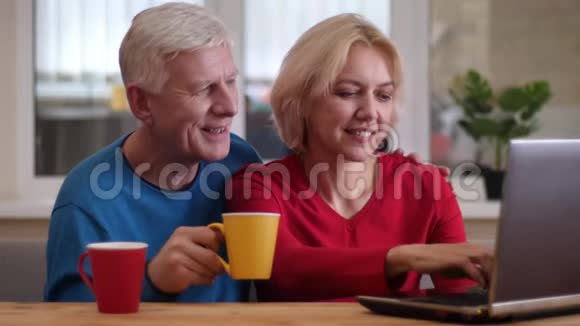 在桌上的笔记本电脑上拍摄高级幸福夫妇网络的特写镜头在舒适的室内喝咖啡视频的预览图