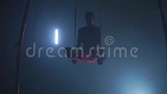 男子体操运动员在黑暗背景下的环上进行倒立和旋转并在缓慢运动中吸烟视频的预览图