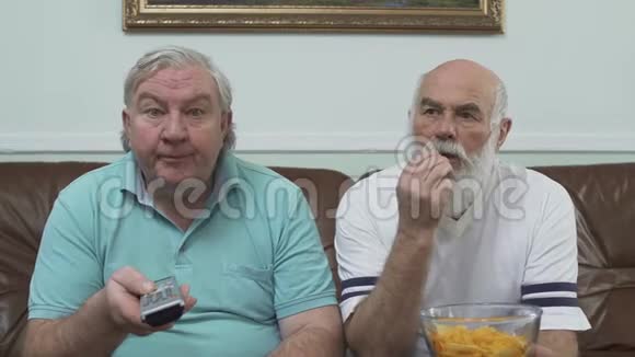 两个成熟的高级男人坐在棕色皮革沙发上看电视的肖像一个拿着薯片的人视频的预览图