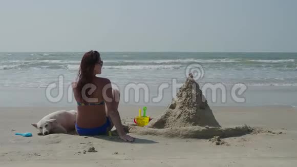 一个穿泳衣的年轻女孩正背着一只白色的狗坐在海滩上那里有一座沙堡4K视频的预览图