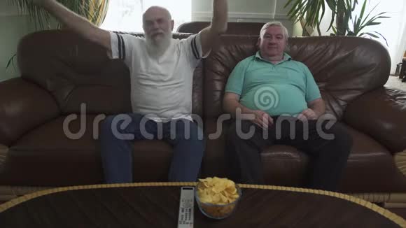 两个成熟的高级男人坐在棕色的皮革沙发上看电视长胡子的情感男人是足球队的球迷这就是视频的预览图
