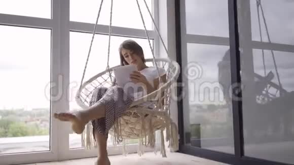 可爱的年轻女人在一张挂着的椅子上摆动同时在大的落地窗上使用她的平板电脑自由职业者视频的预览图