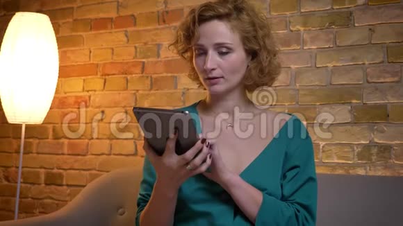 姜黄色卷发的白种人女孩在平板电脑上打字然后在舒适的家里把它的蓝色屏幕显示视频的预览图