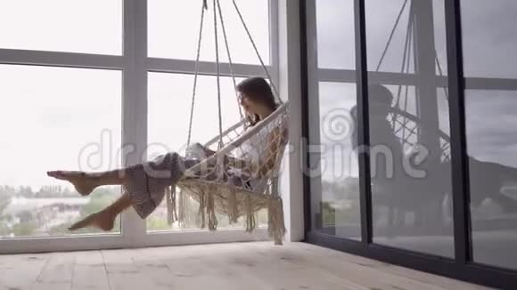 漂亮的年轻女子在一张挂着的椅子上荡秋千在大的落地窗上用平板电脑视频的预览图