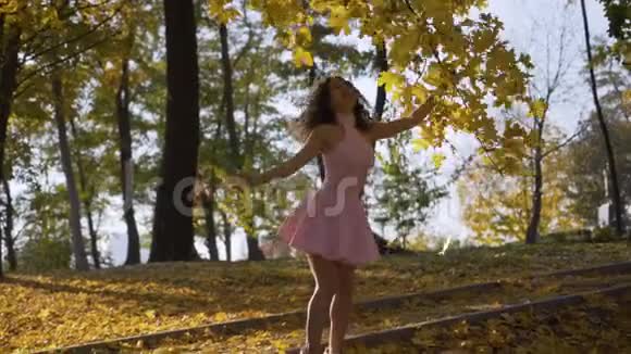 更远的距离公园里有趣的卷曲女孩在金色的树间旋转温暖的秋日柔和的灯光4K视频的预览图
