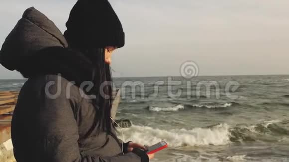 在海边戴耳机的漂亮女孩正在寻找手机上的音乐视频的预览图
