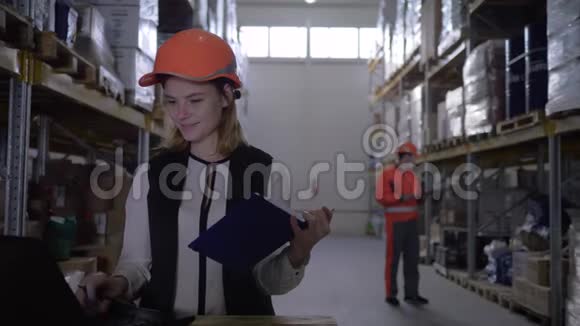戴安全帽的员工女士使用笔记本电脑并在仓库工人的背景下在记事本上做笔记视频的预览图