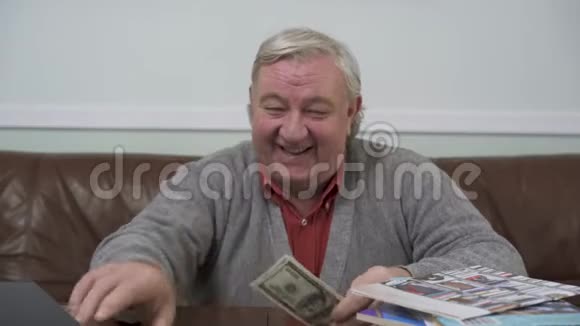 快乐的老人坐在木桌旁手里拿着钱积极的富人展示他的钱视频的预览图