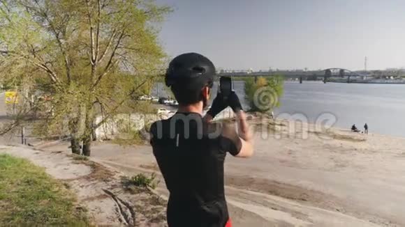 骑自行车的人穿着黑色的衣服头盔和太阳镜拿着他的手机相机拍摄城市河流的照片男性运动员视频的预览图