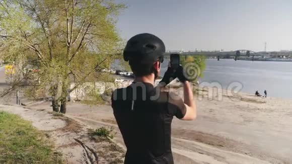 骑自行车的人穿着黑色的衣服头盔和太阳镜拿着他的手机相机拍摄城市河流的照片男性运动员视频的预览图