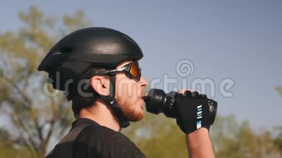 特写三项运动员的肖像戴着黑色头盔和眼镜饮用水大胡子骑自行车的男性从水瓶里喝水视频的预览图