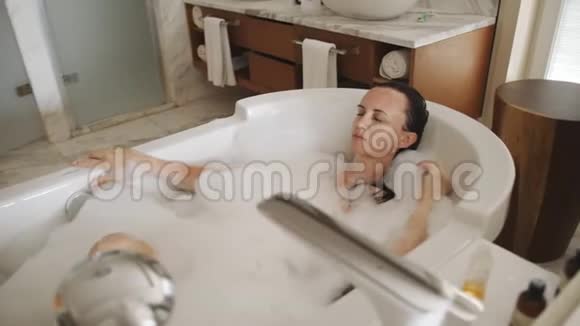 快乐的女孩在浴室里洗了满是泡沫的澡吹肥皂泡沫身体护理视频的预览图