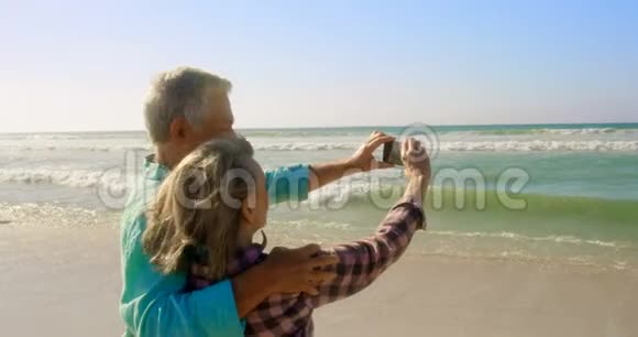 活跃的高加索夫妇在海滩上用手机自拍的后视图视频的预览图