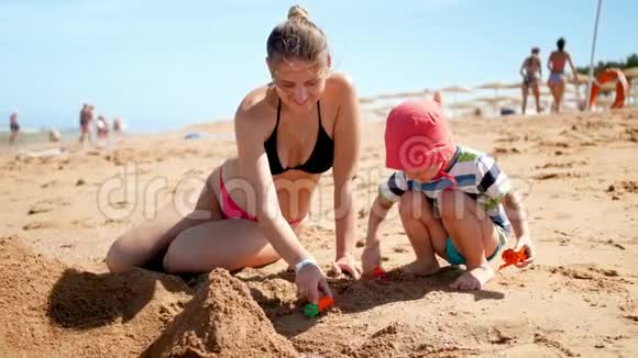 美丽的年轻母亲和她3岁的蹒跚学步的儿子在海滩上玩耍的4K慢镜头家庭放松视频的预览图