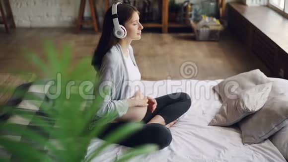美丽的孕妇坐在现代公寓的床上抚摸着她的肚子穿着音乐听着视频的预览图