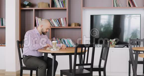 穿着办公室衣服的大胡子秃顶正在看一本书坐在酒店大厅的咖啡馆里喝着一杯卡布奇诺视频的预览图