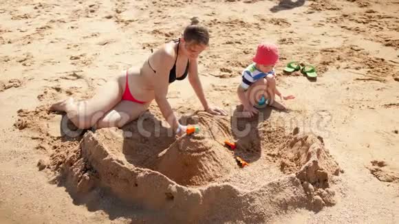 4k镜头拍摄的年轻母亲穿着比基尼和她3岁的儿子在海边建造沙堡视频的预览图