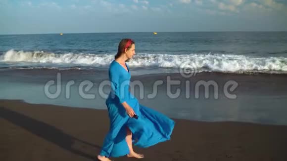 穿着漂亮蓝色连衣裙和太阳镜的女人在海滩上散步快乐地旋转着慢动作视频的预览图