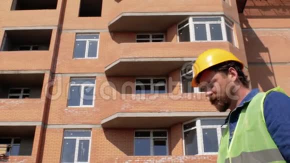 一位戴着黄色头盔留着胡子和胡子的建筑工人走近正在建造的房子讲述了建筑工人的故事视频的预览图