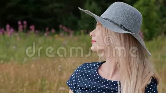 一个年轻迷人的金发女人穿着蓝色的连衣裙和帽子坐在草地上的格子上风吹着头发视频的预览图