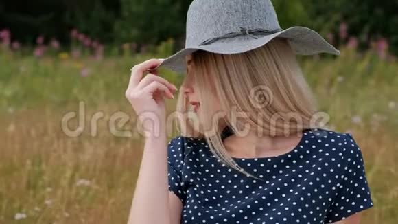 一个年轻迷人的金发女人穿着蓝色的连衣裙和帽子坐在草地上的格子上风吹着头发视频的预览图