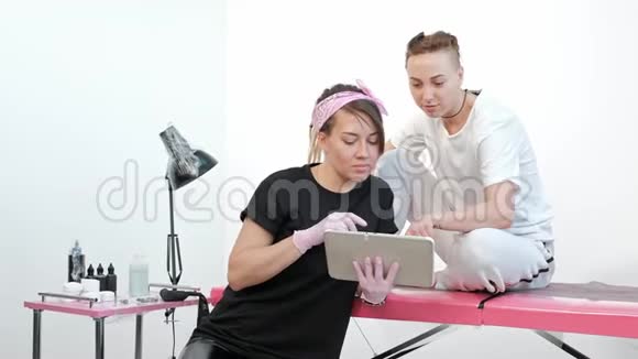 纹身大师女士与客户讨论素描在工作室的平板电脑上展示图片视频的预览图