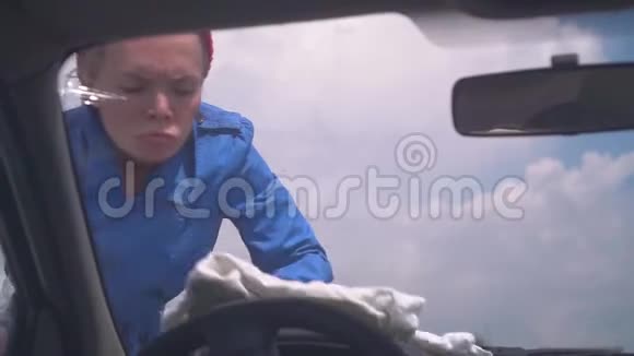 金发女郎玩得很开心穿着蓝色外套戴着红色帽子在户外擦拭汽车的玻璃幽默视频的预览图