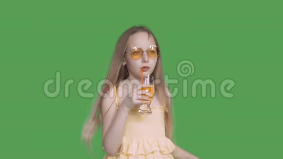 年轻的女孩戴着太阳镜在透明的绿色背景下用吸管从玻璃中喝橙汁少女视频的预览图