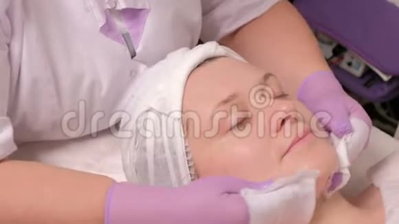一个中年妇女在美容院做手术的画像美容师穿紫色手套的治疗师擦拭视频的预览图