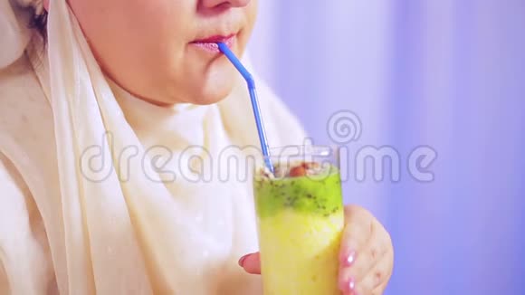 一位年轻的穆斯林妇女戴着一条浅色围巾手里拿着一个杯子手里拿着一块水果冰沙用吸管喝着东西视频的预览图