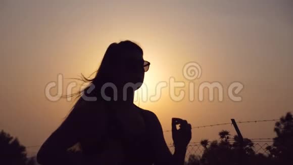 日落时分在乡间小路上慢跑和听音乐的运动女孩剪影戴墨镜的年轻女子视频的预览图