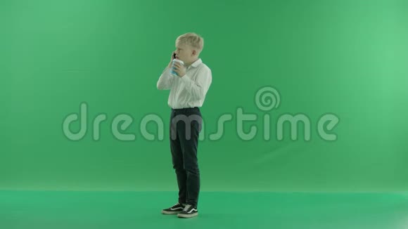一个穿着校服的金发小男孩正拿着一杯咖啡或茶用手机说话他站着视频的预览图