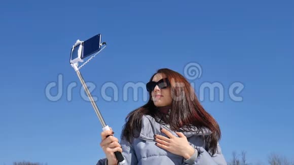 在阳光明媚的公园里戴着太阳镜微笑的年轻女子正在用智能手机和自拍棒拍照视频的预览图
