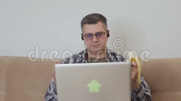 一名中年男子坐在沙发上用笔记本电脑吃香蕉并通过视频链接与对话者沟通视频的预览图