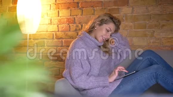在舒适的室内沙发上坐着用平板电脑拍摄白种人成年金发女性的近景拍摄视频的预览图
