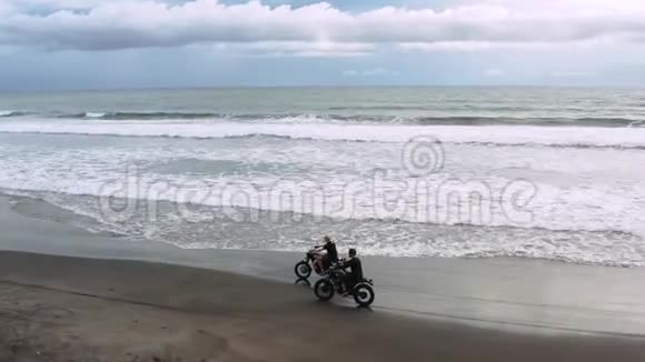 两个英俊的嬉皮士男子骑着现代定制摩托车咖啡馆赛车在黑色沙滩附近的海洋冒险视频的预览图
