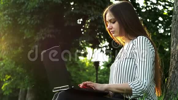 年轻漂亮的女商人在笔记本电脑上打字坐在靠近树的草地上女人在公园里用笔记本视频的预览图