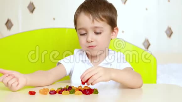 小快乐的孩子坐在桌子旁看着明亮的水果糖果男孩拿着糖果吃了它视频的预览图