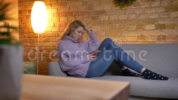 在室内放松的沙发上悠闲地坐着的白种人成年金发女性的特写镜头视频的预览图