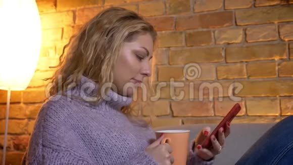 特写侧景拍摄成年白种人金发女性在沙发上悠闲地坐着浏览电话视频的预览图