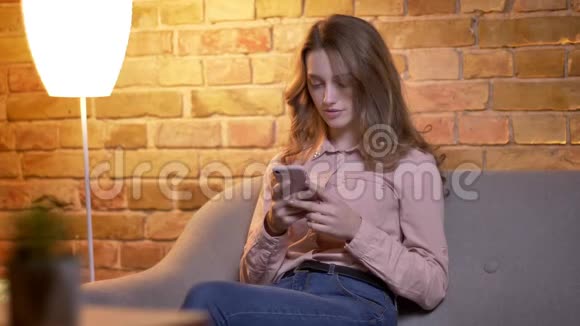 年轻迷人的少女的肖像智能手机在舒适的家中欢乐地对着镜头微笑调整头发视频的预览图