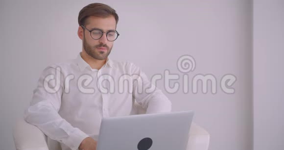 戴着眼镜长着大胡子的白种人成年商人的特写镜头用笔记本电脑看着镜头微笑视频的预览图