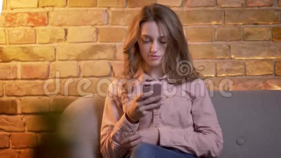 年轻迷人的少女坐在沙发上在舒适的家庭氛围中观看智能手机的肖像视频的预览图