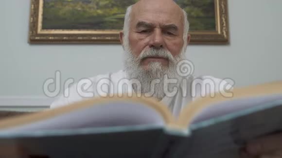 特写肖像体贴的胡须老人坐在木桌上坐在皮革沙发上看书高加索人视频的预览图