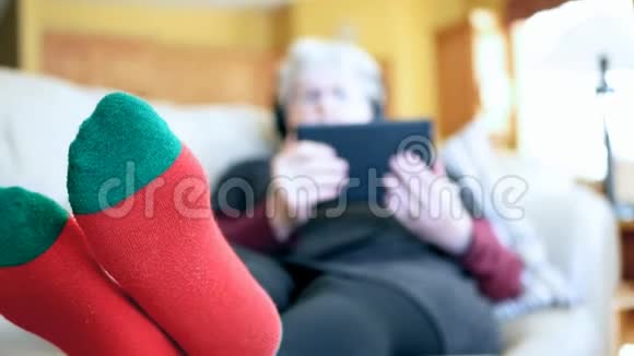 穿着红袜子的老妇人在家里的沙发上用平板电脑和耳机放松祖母阅读和听音乐视频的预览图